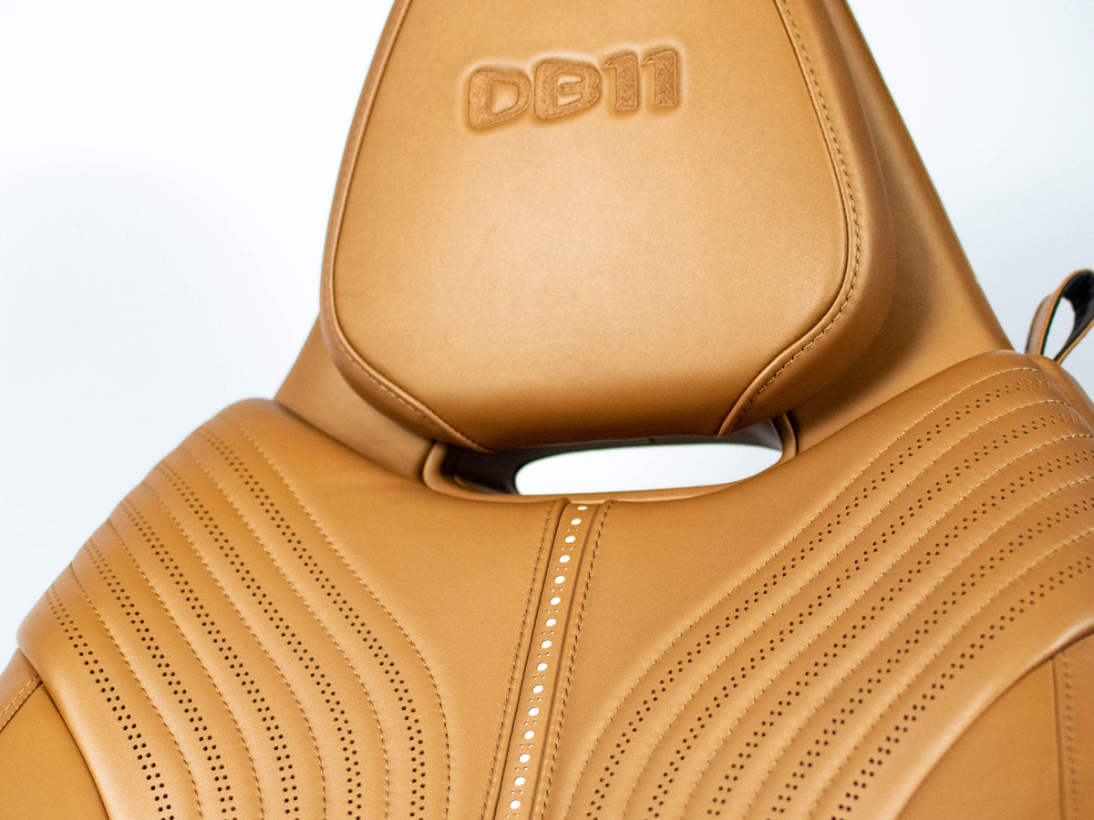 Close-up of a tan leather car seat with a contoured headrest, decorative stitching, and an embossed 'DB11' logo on the headrest.