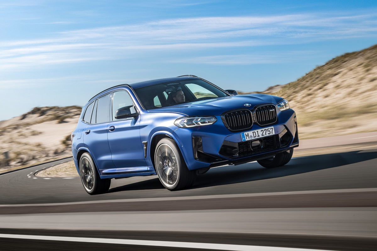 A blue BMW X3 SUV is shown driving on a paved road with sandy hills and a blue sky in the background.