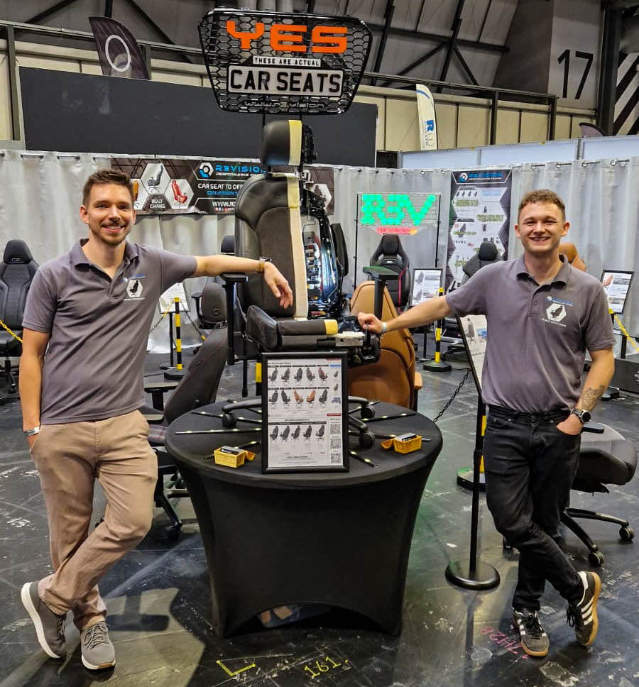 R3VISION team in matching gray polo shirts stand on either side of a black display table showcasing a cut through car seat office chair at an exhibition stall. A large sign above reads 'YES these are CAR SEATS', and other booths and signage are visible in the background.